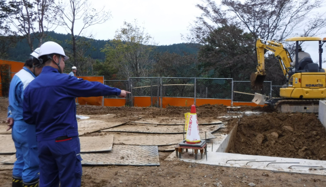 屋外工事現場で工程を確認する様子。奥にユンボ